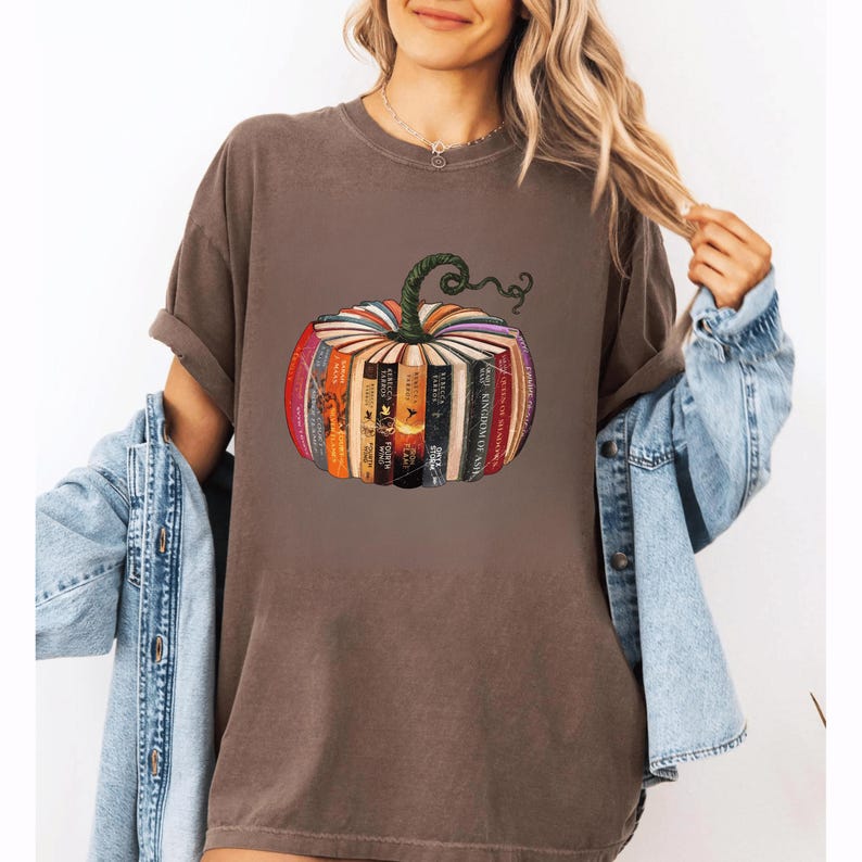 May include: Brown t-shirt featuring a pumpkin design made of books. The pumpkin has a green stem and is composed of various book spines in different colors. The shirt is a relaxed fit and has rolled-up sleeves.