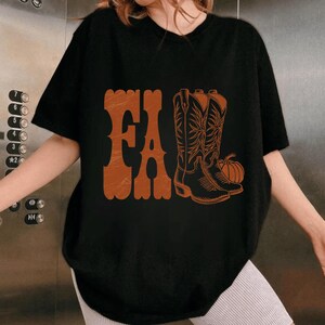 May include: Black t-shirt with the word "FALL" in orange, along with an illustration of cowboy boots and a pumpkin. The design has a vintage, rustic aesthetic, perfect for autumn.