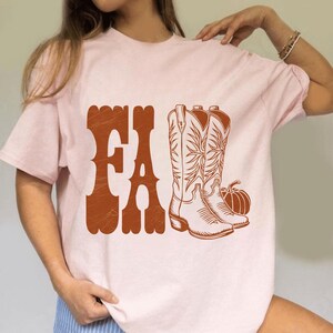 May include: A light pink t-shirt with a brown graphic that says "FA" and features a pair of cowboy boots and a pumpkin. The design has a vintage, rustic aesthetic, perfect for fall.