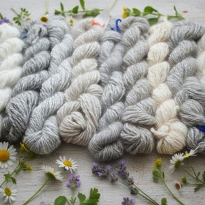 May include: A collection of yarn skeins in various shades of gray and white, arranged in a gradient. The yarn is surrounded by sprigs of flowers and greenery, including daisies and lavender, creating a natural, rustic aesthetic.
