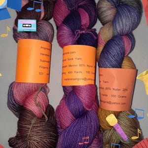 May include: Three skeins of hand-dyed sock yarn in shades of purple, pink, brown, and blue. Each skein is labeled with details including the yarn type, weight, and fiber content. The yarn is presented with a musical theme, featuring musical notes and instruments.