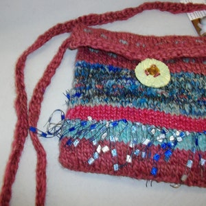 May include: A small knitted handbag with a pink, blue, and gray striped pattern. The bag has a flap with a button closure and a long strap. The bottom of the bag is decorated with a fringe of blue and silver beads.