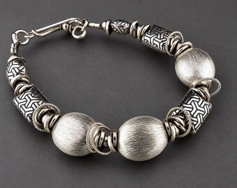 May include: A silver-toned bracelet featuring a series of large, brushed-finish spherical beads alternating with rectangular beads that display a black and silver geometric pattern, accented by smaller silver rings and a hook-style clasp closure.