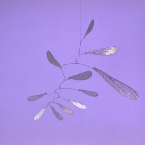 Puede incluir: Una escultura móvil plateada con múltiples alas delicadas y translúcidas que cuelgan de un fino alambre. Las alas están dispuestas en cascada, creando una sensación de movimiento y ligereza.