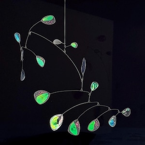 Könnte beinhalten: Eine moderne Mobile-Skulptur mit minimalistischem Design. Das Mobile zeigt schillernde, tropfenförmige Elemente in Grün-, Rosa- und Blautönen, die an dünnen, silbernen Drähten aufgehängt sind. Das Kunstwerk ist vor einem dunklen Hintergrund platziert.