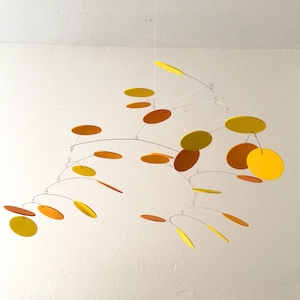 May include: A modern mobile sculpture with a series of colorful, circular shapes suspended from thin wires. The discs are in shades of yellow, orange, and brown. The mobile is hanging from the ceiling against a white background.