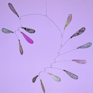 Puede incluir: Móvil moderno con elementos en forma de hojas en varios colores, incluyendo morado, plateado y dorado. Las hojas están suspendidas de finos alambres curvos, creando un diseño dinámico y aéreo sobre un fondo lavanda.