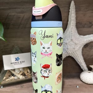 May include: A light green water bottle with a black, pink, and light green lid. The bottle is decorated with various cat faces and the name "Yami". The lid has the text "Lift to Unlock".
