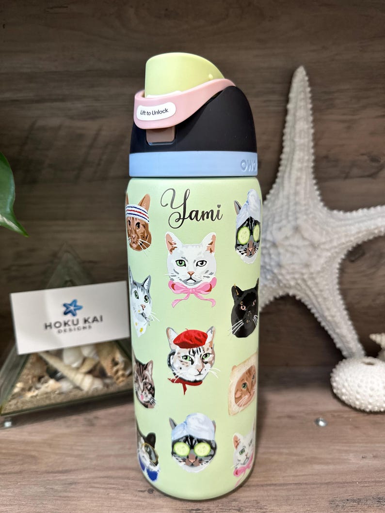 May include: A light green water bottle with a black, pink, and light green lid. The bottle is decorated with various cat faces and the name "Yami". The lid has the text "Lift to Unlock".