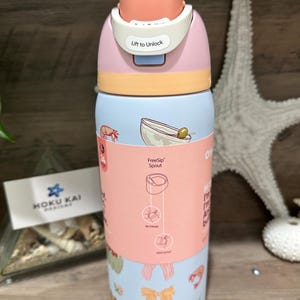 May include: A colorful water bottle with a pink, peach, and light blue design. The top has a peach-colored flip-top lid with the text "Lift to Unlock." The bottle features illustrations of shrimp, olives, and bows. The text "FreeSip Spout" is also visible.