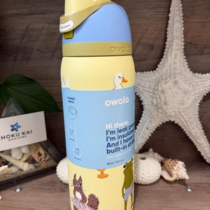Puede incluir: Una botella de agua Owala FreeSip amarilla y azul con una ilustración de pato y ardilla. La botella tiene una pajita incorporada y contiene 945 ml. La tapa es amarilla y azul con un botón para desbloquear.