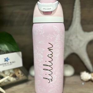 May include: A pink water bottle with a floral design and the name "Lillian" in cursive. The bottle has a white and pink lid with a "Lift to Unlock" label. The bottle is made of stainless steel.