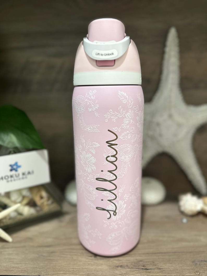 May include: A pink water bottle with a floral design and the name "Lillian" in cursive. The bottle has a white and pink lid with a "Lift to Unlock" label. The bottle is made of stainless steel.