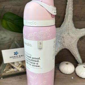 May include: A pink Owala water bottle with a white lid and accents. The bottle has a floral design and the text "owala" on the top. The bottle is insulated, leak-proof, and has a built-in straw. The bottle is 9 inches tall.