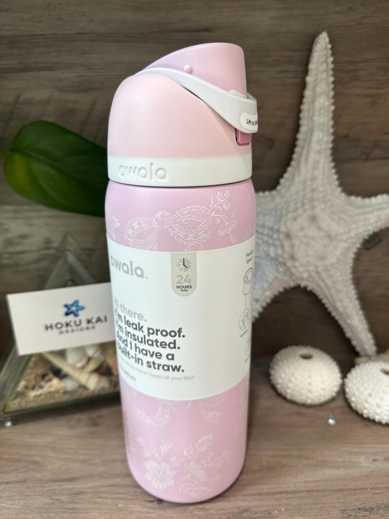 May include: A pink Owala water bottle with a white lid and accents. The bottle has a floral design and the text "owala" on the top. The bottle is insulated, leak-proof, and has a built-in straw. The bottle is 9 inches tall.