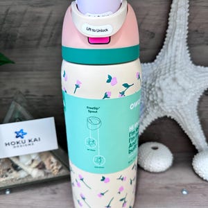 May include: A floral-patterned water bottle with a pink and teal lid. The bottle has a light beige base with pink and teal floral designs. The lid has the text "Lift to Unlock". The bottle is 9 inches tall and holds 17 ounces.