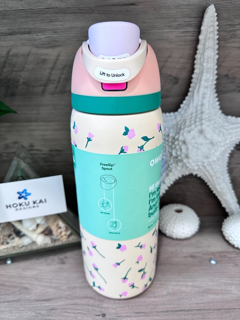 May include: A floral-patterned water bottle with a pink and teal lid. The bottle has a light beige base with pink and teal floral designs. The lid has the text "Lift to Unlock". The bottle is 9 inches tall and holds 17 ounces.