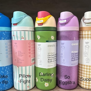 Puede incluir: Cinco botellas de agua aisladas Owala FreeSip en varios colores y patrones. Las botellas tienen tapas abatibles y están etiquetadas con frases como "Take a Dip" y "Suppertime". Cada botella mide aproximadamente 769 ml.