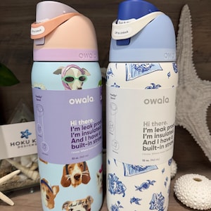 Puede incluir: Dos botellas de agua aisladas Owala. Una es azul claro con ilustraciones de perros y el texto "Hi there. I'm leak proof. I'm insulated. And I have a built-in straw." La otra es blanca con diseños florales azules.