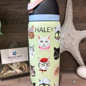 May include: A light green Owala water bottle featuring various cat faces with accessories. The name "HALEY" is printed in white. The bottle has a black, light blue, pink, and yellow lid. A Hoku Kai Designs business card is visible.