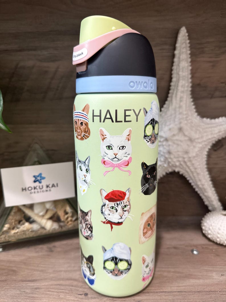 May include: A light green Owala water bottle featuring various cat faces with accessories. The name "HALEY" is printed in white. The bottle has a black, light blue, pink, and yellow lid. A Hoku Kai Designs business card is visible.