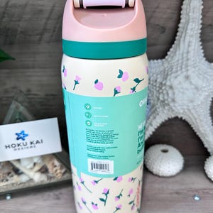 May include: A floral-patterned water bottle with a light pink lid and teal accents. The bottle has a cream-colored base with pink flower designs and teal text. The bottle has the text "Owoks" and "HOKU KAI DESIGNS".