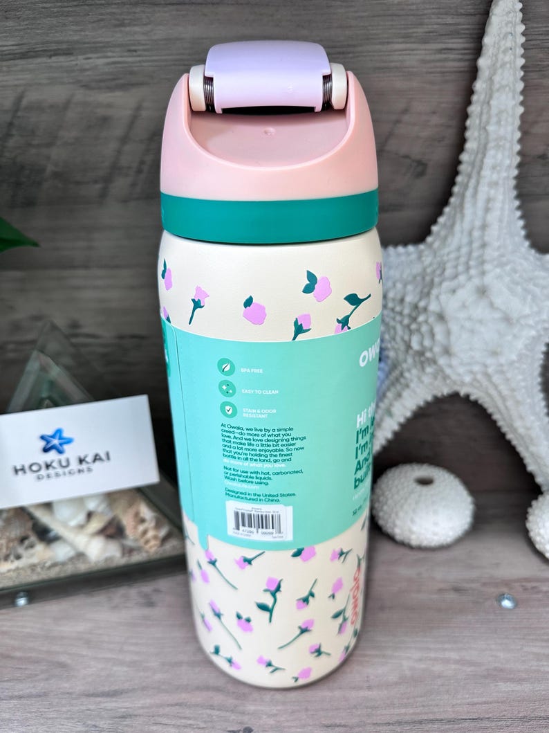 May include: A floral-patterned water bottle with a light pink lid and teal accents. The bottle has a cream-colored base with pink flower designs and teal text. The bottle has the text "Owoks" and "HOKU KAI DESIGNS".