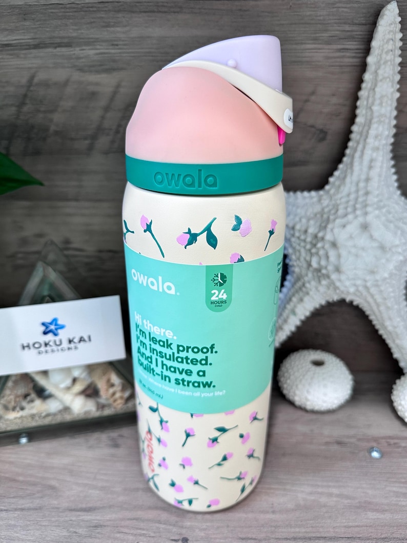 May include: A floral-patterned Owala water bottle with a pink and teal lid. The bottle has a cream background with pink flower illustrations and the text "owala." The bottle also has the text "Hi there. I'm leak proof. I'm insulated. And I have a built-in straw."