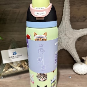 May include: A light green water bottle with a black, pink, and yellow lid. The bottle features cat illustrations and the text "FreeSip Spout". The lid has the text "Push Button" and "Lift to Unlock".