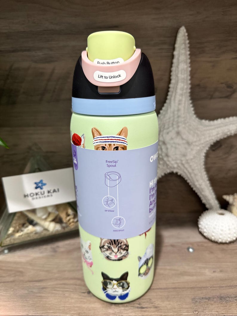 May include: A light green water bottle with a black, pink, and yellow lid. The bottle features cat illustrations and the text "FreeSip Spout". The lid has the text "Push Button" and "Lift to Unlock".
