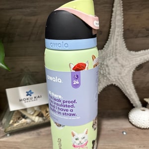 May include: A green Owala water bottle with a black lid, light blue accents, and a pink handle. The bottle features cat illustrations and text that reads, "Hi there. I'm leak proof. I'm insulated. And I have a built-in straw."