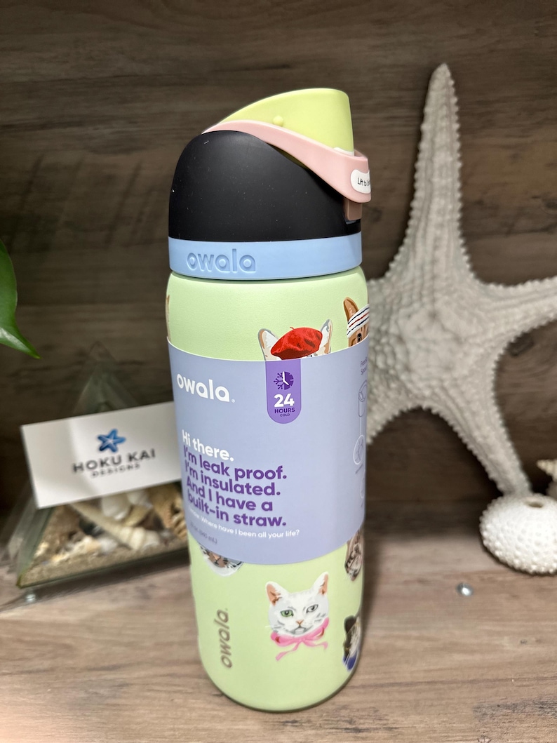 May include: A green Owala water bottle with a black lid, light blue accents, and a pink handle. The bottle features cat illustrations and text that reads, "Hi there. I'm leak proof. I'm insulated. And I have a built-in straw."