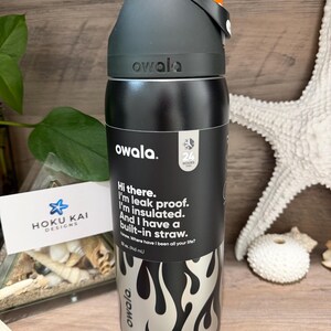 May include: A black and silver Owala water bottle with an orange cap. The bottle has a flame design and text that reads, "Hi there. I'm leak proof. I'm insulated. And I have a built-in straw." It holds 32 oz (945 mL).