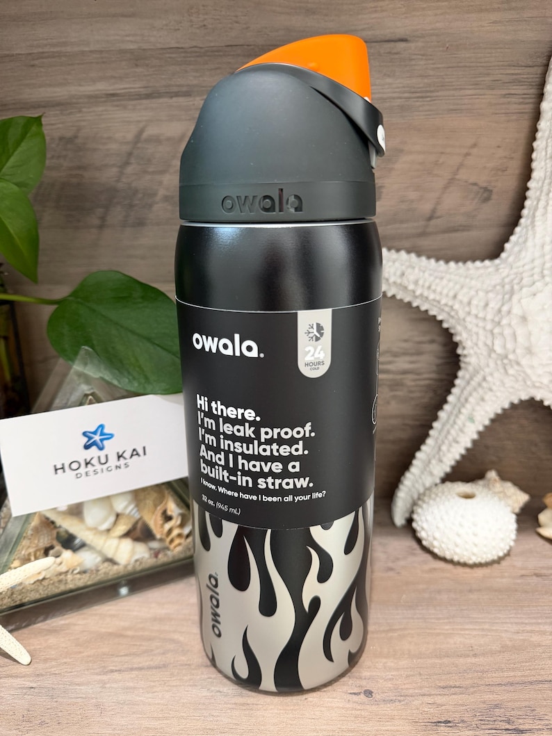 May include: A black and silver Owala water bottle with an orange cap. The bottle has a flame design and text that reads, "Hi there. I'm leak proof. I'm insulated. And I have a built-in straw." It holds 32 oz (945 mL).