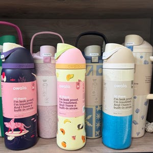 Puede incluir: Una colección de botellas de agua aisladas Owala en varios colores, incluyendo rosa, azul, verde y beige. Cada botella tiene una pajita incorporada y el texto: "Hi there. I'm leak proof. I'm insulated. And I have a built-in straw."