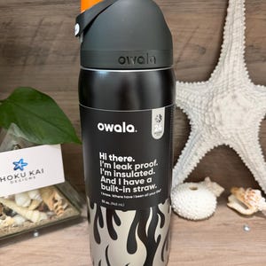 May include: A black and silver Owala water bottle with an orange lid. The bottle has a flame design and text that reads, "Hi there. I'm leak proof. I'm insulated. And I have a built-in straw." It holds 32 oz. (945 mL).