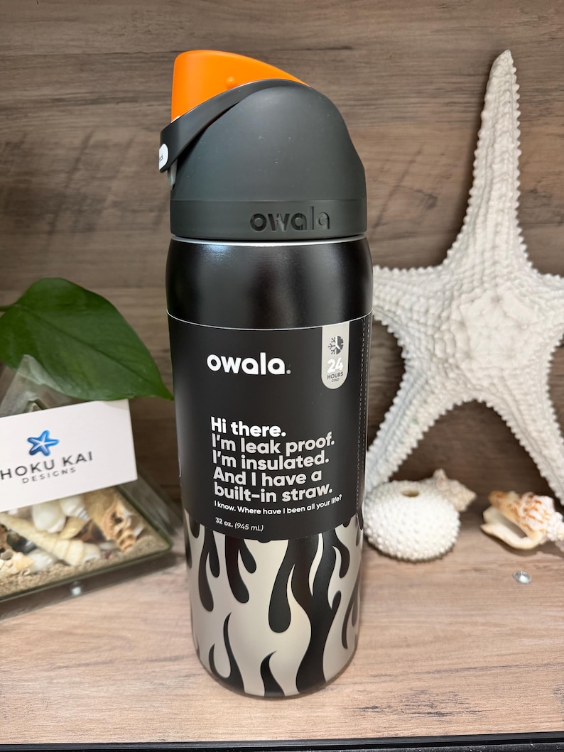 May include: A black and silver Owala water bottle with an orange lid. The bottle has a flame design and text that reads, "Hi there. I'm leak proof. I'm insulated. And I have a built-in straw." It holds 32 oz. (945 mL).