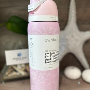 May include: A pink Owala insulated water bottle with a floral design. The bottle has a white lid and a built-in straw. The text on the bottle says, "Hi there. I'm leak proof, I'm insulated, and I have a built-in straw." It holds 32 oz.