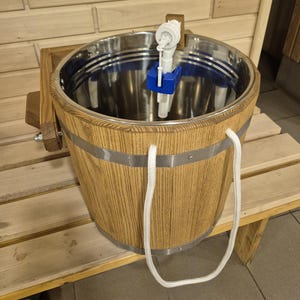 Könnte beinhalten: Ein Saunaeimer aus Holz mit Edelstahl-Innenraum und weißem Seilgriff. Der Eimer hat Metallbänder und einen blau-weißen Schwimmer. Der Eimer steht auf einer Holzbank.
