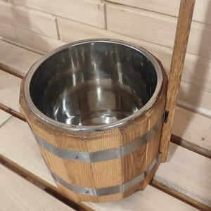 Könnte beinhalten: Saunaboot aus Holz mit Edelstahl-Innenraum und langem Holzgriff. Der Eimer besteht aus hellbraunem Holz und wird von Metallbändern zusammengehalten. Der Griff ist an der Seite des Eimers befestigt.