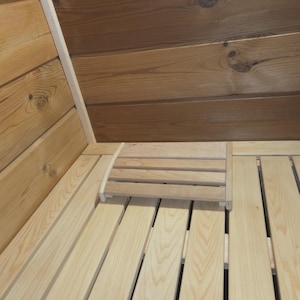 May include: Interior view of a sauna with light-colored wooden planks on the floor and walls. A small, slatted wooden seat is positioned on the floor. The ceiling is made of darker wood.