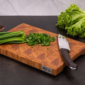 Op de afbeelding: Een houten snijplank met gehakte groene uien en een klein mes. Verse sla en een houten lepel met peperkorrels zijn ook aanwezig. De plank heeft een uniek patroon.