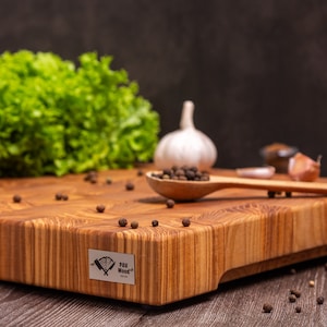 Può includere: Tagliere in legno marrone chiaro di forma rettangolare. Un cucchiaio di legno contiene grani di pepe. Altri ingredienti sono lattuga, pomodori e aglio. Il tagliere ha un'etichetta metallica con la scritta "PAV Wood".