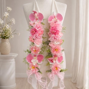 May include: A floral lei displayed on a mannequin. The lei features pink orchids, dahlias, and other flowers, accented with green foliage and white ribbons. The lei is strung on a white beaded necklace.