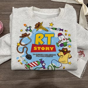 May include: A light gray crewneck sweatshirt featuring a colorful graphic print with the text RT STORY TO INFINITY AND BEYOND FOR OUR PATIENTS. The design includes illustrations of a stethoscope, syringe, oxygen tank, and medical items.