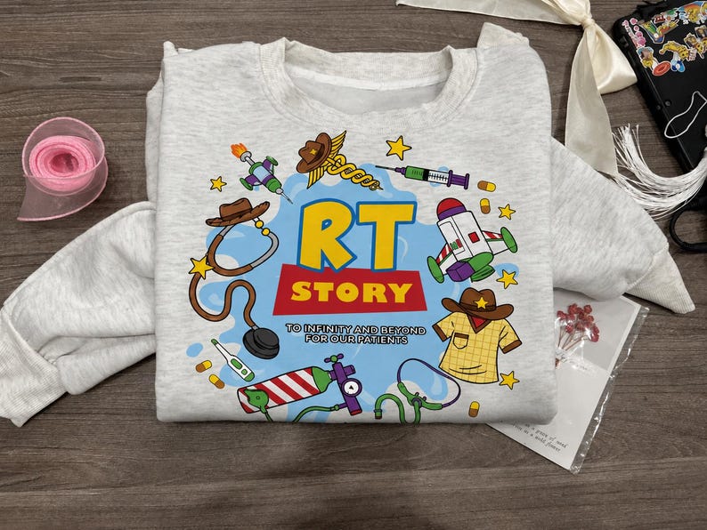May include: A light gray crewneck sweatshirt featuring a colorful graphic print with the text RT STORY TO INFINITY AND BEYOND FOR OUR PATIENTS. The design includes illustrations of a stethoscope, syringe, oxygen tank, and medical items.