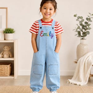Könnte beinhalten: Eine hellblaue Jeans-Latzhose mit dem Namen Emily in Regenbogenfarben auf der Brust gestickt, kombiniert mit einem kurzärmeligen T-Shirt mit roten und weißen Querstreifen sowie weißen Canvas-Sneakern mit Schnürsenkeln.