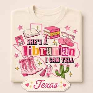 May include: A cream-colored t-shirt featuring a pink outline of the state of Texas. The graphic includes a stack of books labeled Fiction, Mystery, and Romance, a library card, pink cowboy boots, a pink cowboy hat, glasses, and a cactus.