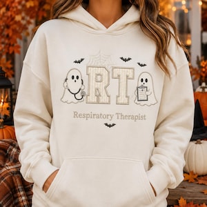 May include: A cream-colored hooded sweatshirt featuring an embroidered design with two ghosts, one holding a stethoscope and the other a clipboard, flanking the letters RT. Above is a spiderweb and bats, with the text Respiratory Therapist below.