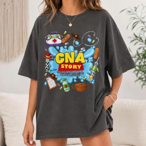 May include: A charcoal gray t-shirt featuring a colorful graphic with the text CNA STORY TO INFINITY AND BEYOND FOR OUR PATIENTS. The design includes medical icons like a first aid kit, stethoscope, syringe, mask, and pills with cowboy motifs.